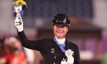 Isabell Werth smiles and waves in celebration while holding flowers in her right hand and a silver medal in her left