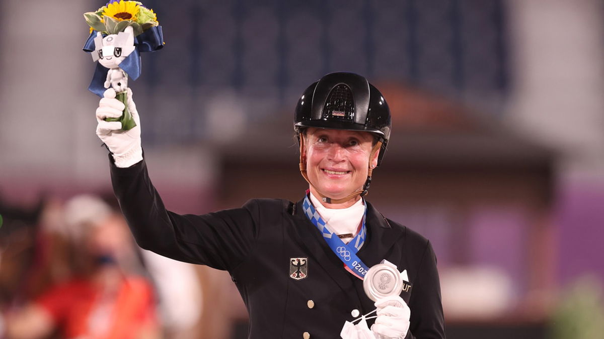 Isabell Werth smiles and waves in celebration while holding flowers in her right hand and a silver medal in her left