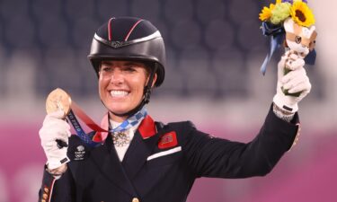 Charlotte Dujardin smiles and holds a bronze medal in her right hand and raises flowers in her left hand